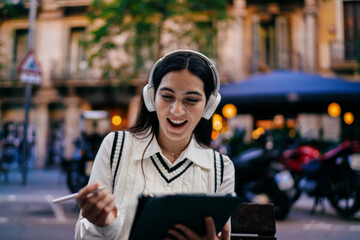 Woman wearing headphones sits outdoors drawing on tablet with stylus, enjoying portable digital art creation — blending street mood and mobile tech for expressive design.