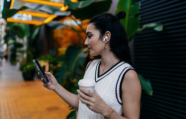 Young woman walks outdoors holding smartphone and coffee, wearing wireless earbuds, reflecting mobile tech use and multitasking on the go in casual urban lifestyle.