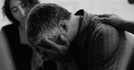 Man sobs during therapy session, burying face in hands while others gently place hands on his back and shoulder in gesture of deep emotional support and empathy