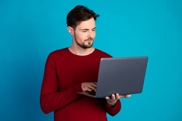 Young man with laptop wearing red sweater stands against blue background looking at the screen in a casual stylish pose