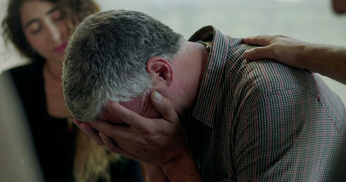 Man breaks down in therapy session, covering face with hands as group members offer comfort with hand on shoulder in scene of deep emotional release and support - Powered by Adobe