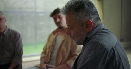 Man speaking earnestly during group therapy session, expressing emotional burden with open hands as peers listen with compassion in supportive setting