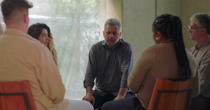 Man speaks during group therapy session, leaning forward while engaging with others in supportive circle inside quiet room filled with natural light and emotional focus