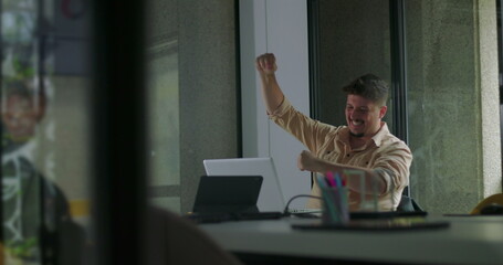 Man celebrates success with excited arm gesture at office desk, smiling at laptop after positive results in modern workplace