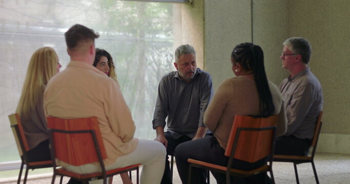 Man speaks during group therapy session, leaning forward while engaging with others in supportive circle inside quiet room filled with natural light and emotional focus