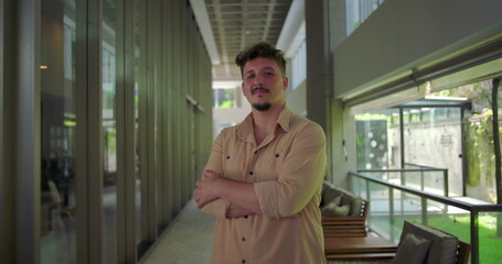 Man standing in office hallway with arms crossed, confident expression, assertive posture in modern workplace, strength and leadership presence