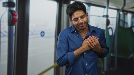 Young hispanic man with hands clasped over chest on airport shuttle bus, wearing glasses and a blue shirt, eyes closed in a grateful smile; gratitude reflection.