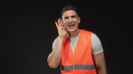 Man in orange safety vest holds hand to ear, straining to hear in studio with black background; frustration.