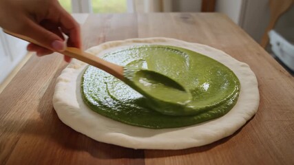 Spreading vibrant green pesto sauce on raw pizza dough on rustic wooden surface in bright kitchen