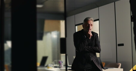 Mature businessman reflecting silently during office meeting with hand on chin and distant gaze in modern corporate boardroom