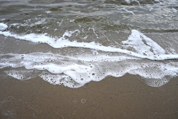 High-resolution abstract texture of the water's edge. The smooth sand meets the dynamic foam and dark water, creating a moody, tranquil natural background for travel or environmental themes.