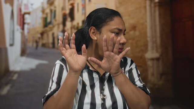 Referee woman holds hands raised near face with whistle hanging from striped jersey on sunlit old town street; caution.