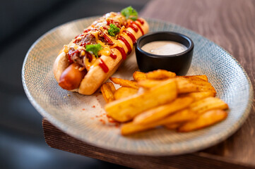 Gourmet hot dog with chili, mustard, ketchup, and herbs, served with crispy potato wedges and dipping sauce on a rustic plate