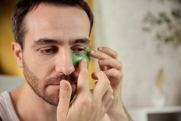 Man placing green under-eye patches for refreshing skincare treatment. Concept of wellness routines, beauty branding, grooming education and fatigue-relief promotional visuals.