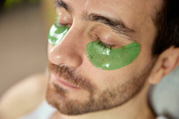 Man relaxing with hydrating green under-eye patches during skincare break. Concept of wellness platforms, beauty branding, self-care visuals and fatigue-recovery treatments.