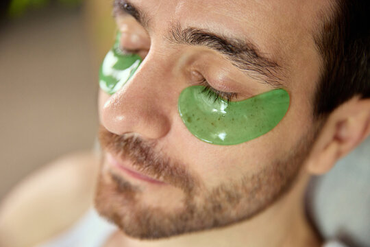 Man relaxing with hydrating green under-eye patches during skincare break. Concept of wellness platforms, beauty branding, self-care visuals and fatigue-recovery treatments.