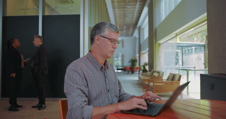 Fototapeta premium Middle aged man resuming work on laptop at shared office space, focused and typing with coworkers talking in background, overcoming concerns and staying productive