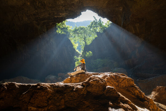 Woman in Yellow Hat Sitting on Boulder Gazing at Sunbeams Streaming Into Tiankeng Sinkhole in Guangxi China