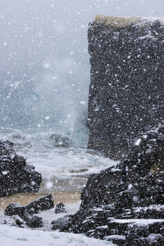 View of a cold winter scene where the fierce ocean waves crash against the rugged, snow-covered cliffs under a heavy snowfall, Snaefellsnes, Eyja- og Miklaholtshreppur, Iceland.