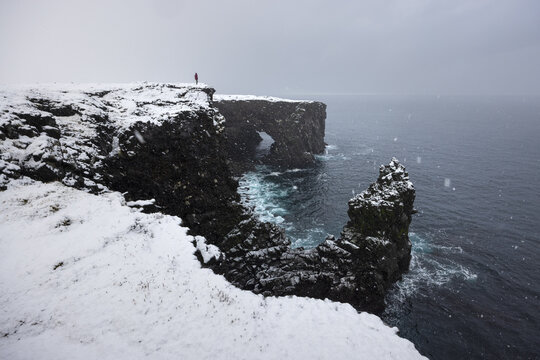 View of snow-dusted cliffs meet the turbulent sea, a lone figure stands against the arctic wind, embracing the stark beauty, Snaefellsnes, Eyja- og Miklaholtshreppur, Iceland.