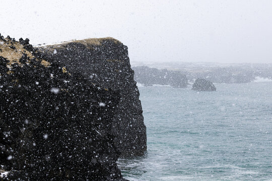 View of a dramatic black lava cliff face meeting the turbulent, teal ocean under a sky of falling snow, Snaefellsnes, Eyja- og Miklaholtshreppur, Iceland.