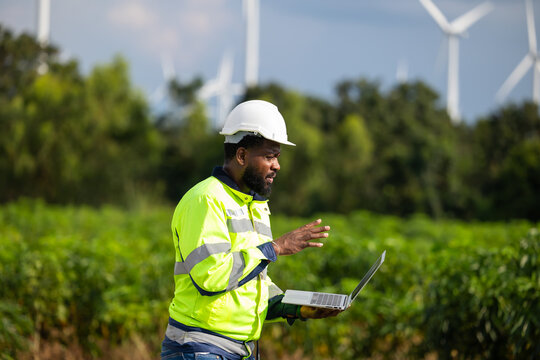 laptop computer. African Engineer man working in field with wind turbines background, Windmill. environmental renewable clean energy. Wind power generation. Windmill engineer inspection progress - Powered by Adobe