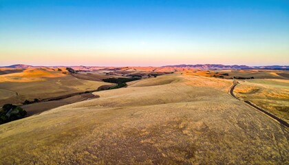 Aerial view of rolling hills covered in dry grass with a road winding through the landscape at sunset. The sky is a gradient of blue and orange.