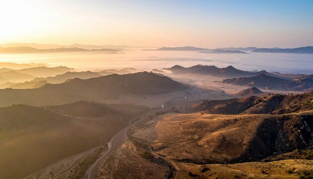 Aerial view of a mountain range with fog in the valley during sunrise, creating a hazy and atmospheric landscape.