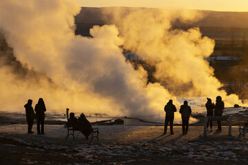 View of rising steam from a geyser illuminated by the golden light of the setting sun, with tourists watching in awe, Golden Circle, Rangarbing eystra, Iceland.