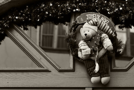 Alsace, France. Christmas wreath decoration on house door with teddy bear holding Christmas tree. "Joyeux Noel" greeting ("Merry Christmas" in French). Christmas holiday celebration background. Sepia 