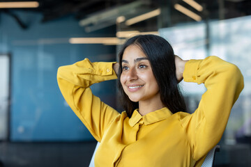 Close-up photo of a young Indian woman in a yellow shirt sitting in an office chair, her hands behind her head, and resting contentedly