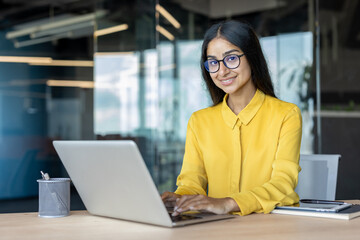 Portrait of a young smiling Indian woman in glasses and yellow shirt sitting at an office desk, working on a laptop and looking at the camera