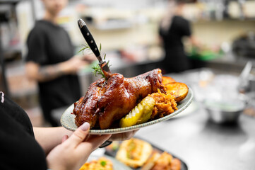 person's hands holding a plate with a large, golden-brown roasted pork knuckle, served with baked potatoes and pickled vegetables in a restaurant kitchen setting.
