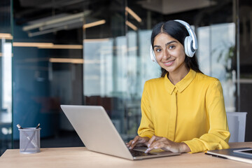 Portrait of a smiling Indian woman in headphones working in the office at a laptop and looking at the camera