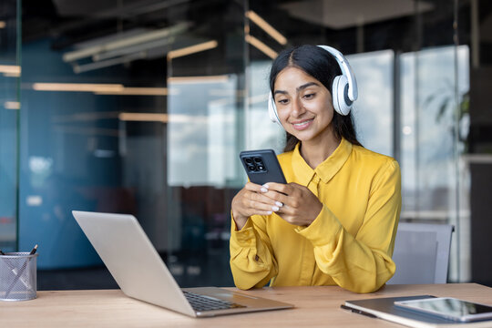 Smiling young Indian woman sitting at office desk wearing headphones and using mobile phone