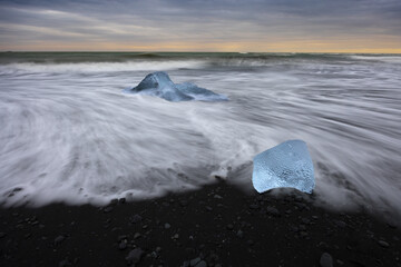 View of glacial ice, shimmering blue against the black sand, kissed by the foamy embrace of the Atlantic waves under a moody sky, Diamond Beach, Sveitarfelagid Hornafjordur, Iceland.