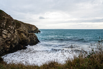 View of the rugged coastline where the dark rocks meet the turquoise sea under a cloudy sky, waves crashing against the shore, St. Davids, Wales, United Kingdom.