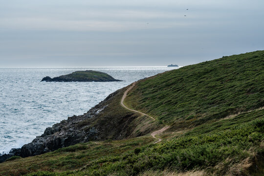 View of a winding trail beckons along a verdant, grassy headland, where the rugged cliffs meet the vast, shimmering sea under a serene sky, St. Davids, Wales, United Kingdom.