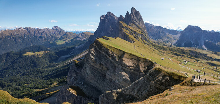 View of jagged peaks pierce the blue sky, casting shadows on the green slopes where tiny figures walk, a vibrant contrast of rock and nature, Ortisei, Trentino-South Tyrol, Italy.