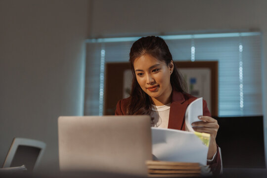 Young Asian businesswoman managing paperwork and working on a laptop at her office desk, demonstrating multitasking and professional efficiency