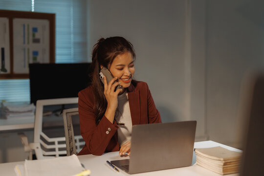 Asian businesswoman having a phone conversation while using a laptop in a dimly lit office, managing tasks efficiently