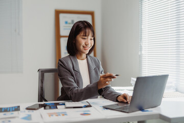 Asian businesswoman working on laptop, smiling while reviewing financial reports and charts in a modern office setup