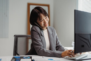 Young Asian businesswoman managing work, talking on a smartphone and typing on a computer keyboard in a modern office