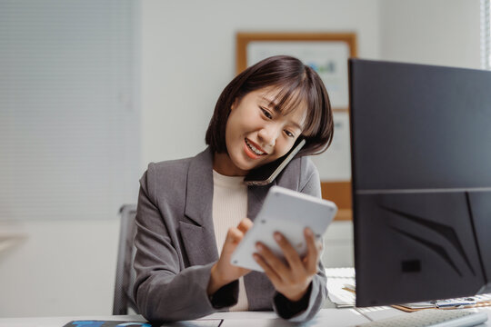 Asian businesswoman smiling, working in an office, holding a calculator and talking on a mobile phone, demonstrating efficiency