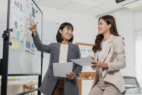 Young happy businesswomen collaborating in an office, reviewing charts and data on a whiteboard during a team meeting - Powered by Adobe