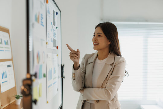 Asian businesswoman analyzing charts and graphs on a presentation board at office meeting, explaining business strategy and planning trends - Powered by Adobe