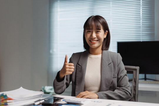 Asian businesswoman smiling and giving a thumbs up gesture, sitting at her office desk, expressing positivity and success