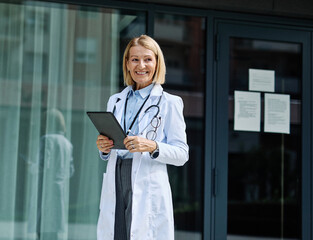 Portrait of a mature female woman doctor or nurse and health care worker holding a cup of coffee and using a tablet computer in front of hospital or clinic outside