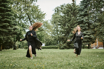 Two young women running joyfully in green park with tall trees