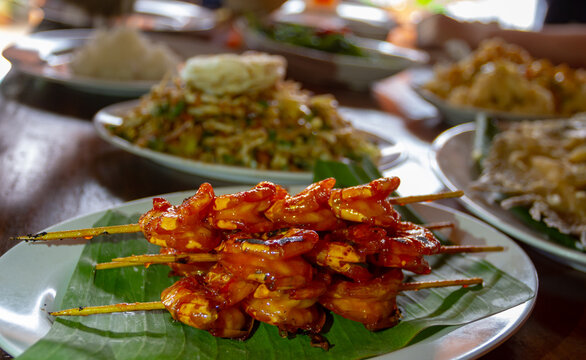 A plate of Sate Udang, Indonesian grilled shrimp skewers glazed with a sweet and spicy sauce. The Penaeus prawns are served on a banana leaf, showcasing char marks and a glossy texture on a wooden tab